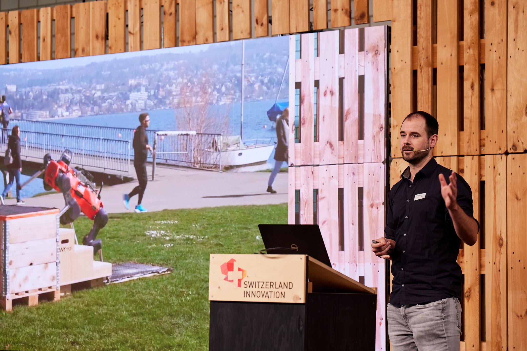 Marco Hutter steht auf der Bühne im Event Hangar vom Innovationspark Zürich  und  präsentiert den roten, vierbeinigen, autonomen Roboter ANYmal von ANYbotics