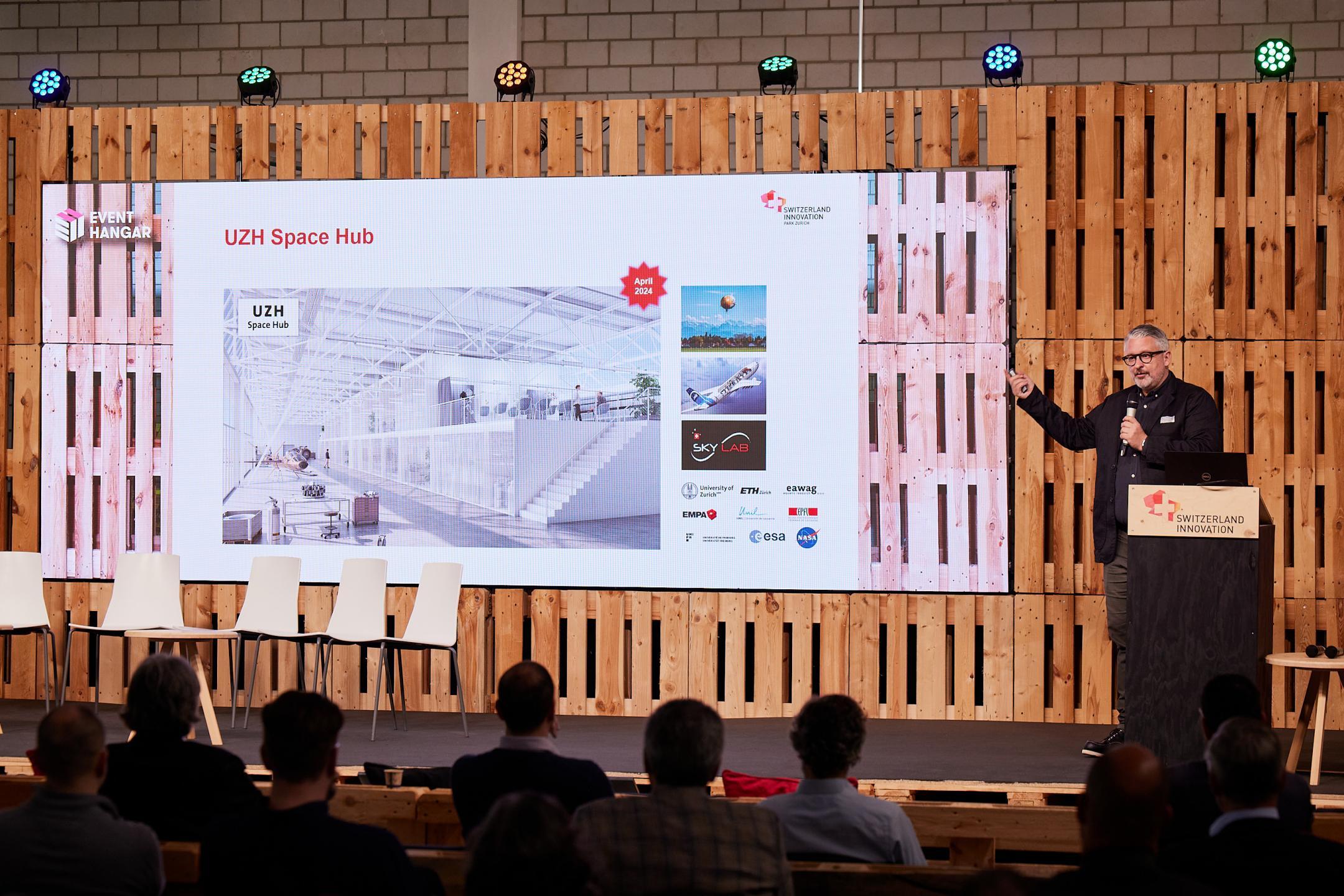 Martin Sturzenegger steht auf der Bühne des Event Hangar im Innovationspark Zürich und präsentiert den UZH Space Hub 