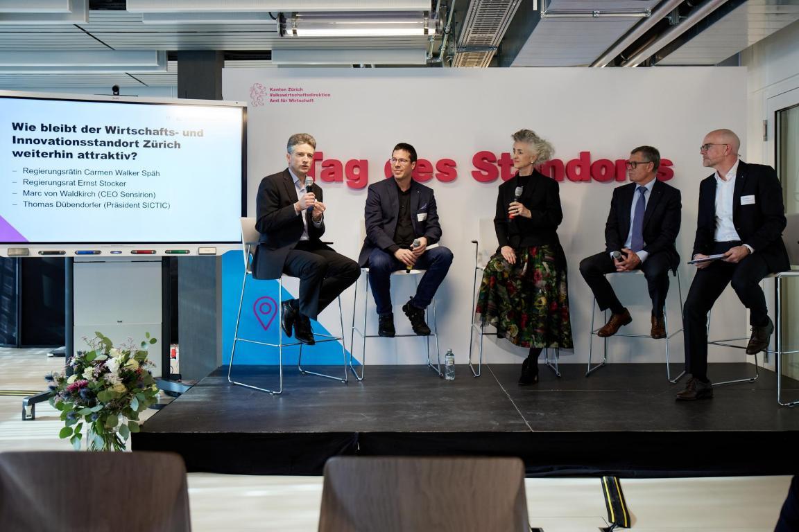 Fünf Personen sitzen auf einem Stuhl und diskutieren miteinander. Zu sehen ist eine Tafel mit der Aufschrift «Wie bleibt der Wirtschafts- und Innovationstandort Zürich weiterhin attraktiv?»