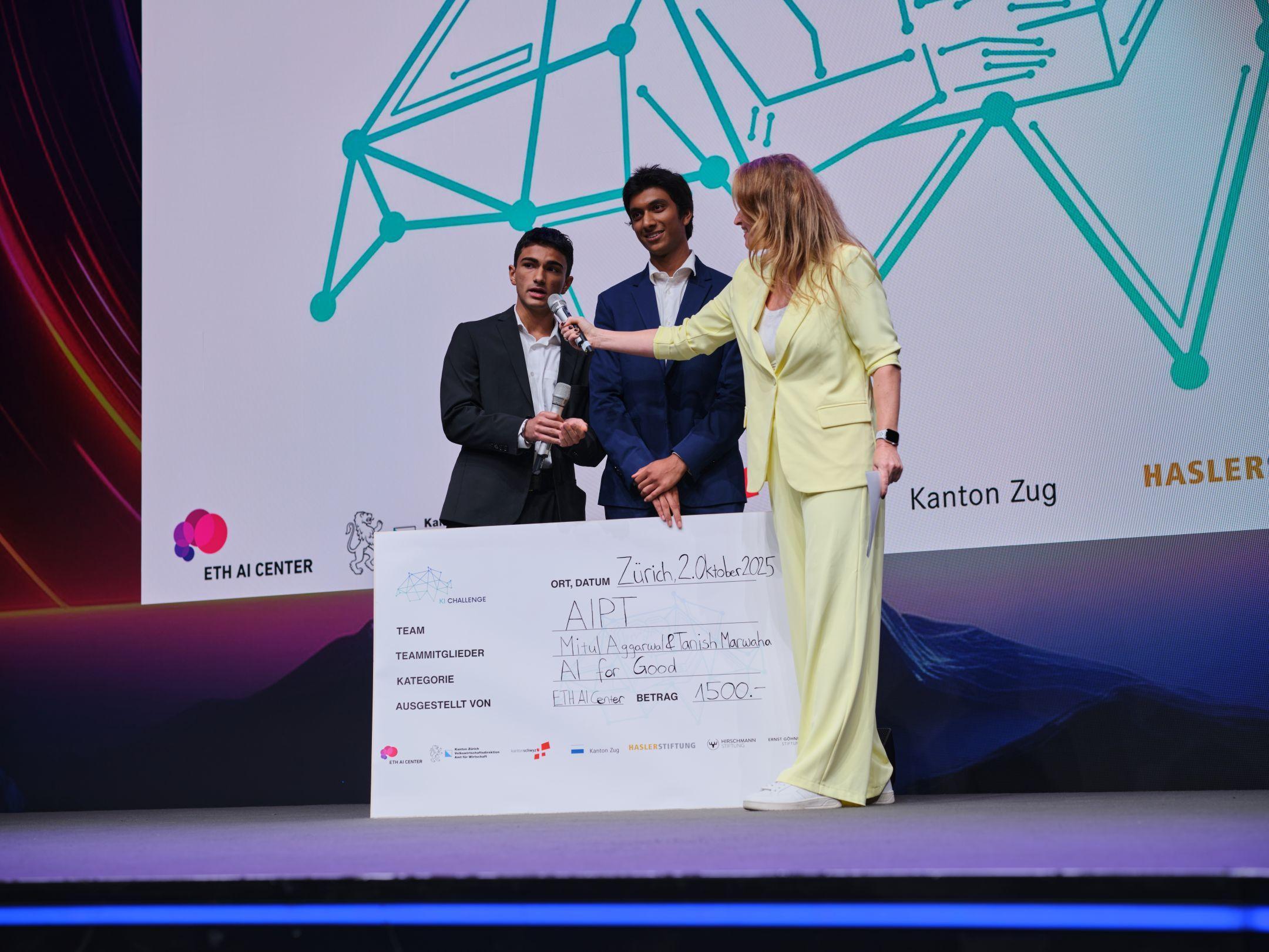 Zwei junge Männer stehen mit Moderatorin Kathrin Hönegger auf der Bühne und präsentieren ihren Siegerscheck der KI Challenge 2025 für das Projekt AIPT.