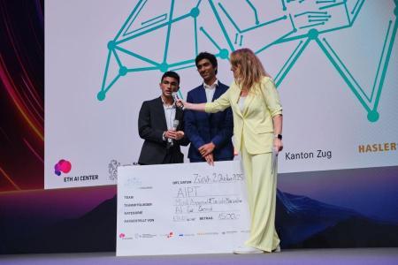 Zwei junge Männer stehen mit Moderatorin Kathrin Hönegger auf der Bühne und präsentieren ihren Siegerscheck der KI Challenge 2025 für das Projekt AIPT.