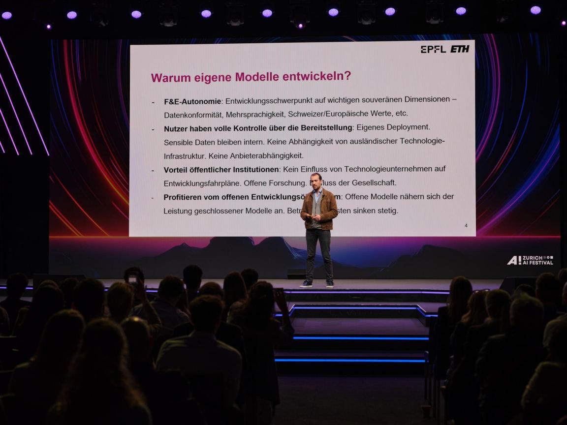 Imanol Schlag präsentiert auf der Bühne des Zurich AI Festivals eine Folie über die Vorteile eigener KI-Modelle vor einem aufmerksam lauschenden Publikum.