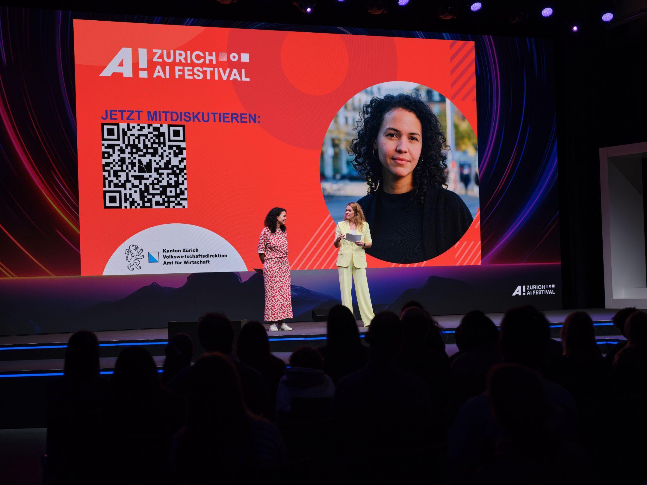 Nathalie Klauser und Moderatorin Kathrin Hönegger stehen auf der Bühne des Zurich AI Festivals vor einer grossen Leinwand mit QR-Code und Festival-Logo.