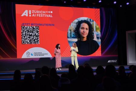 Nathalie Klauser und Moderatorin Kathrin Hönegger stehen auf der Bühne des Zurich AI Festivals vor einer grossen Leinwand mit QR-Code und Festival-Logo.
