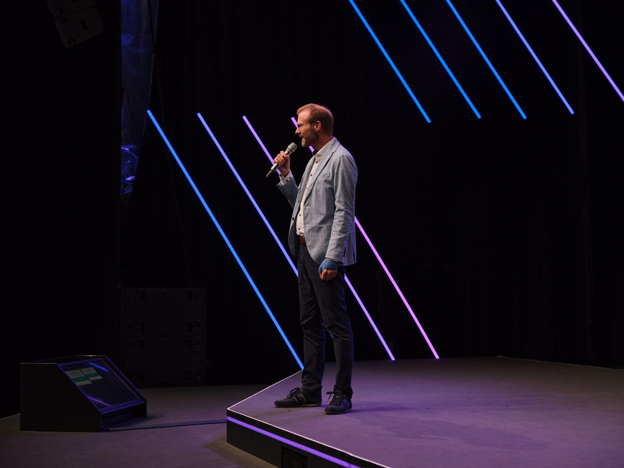 Ein Mann spricht mit Mikrofon auf der Bühne des Zurich AI Festivals, beleuchtet von violetten Lichtstreifen im Hintergrund.