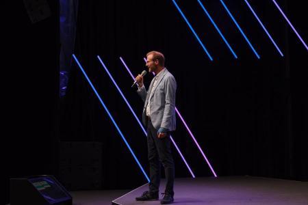 Ein Mann spricht mit Mikrofon auf der Bühne des Zurich AI Festivals, beleuchtet von violetten Lichtstreifen im Hintergrund.