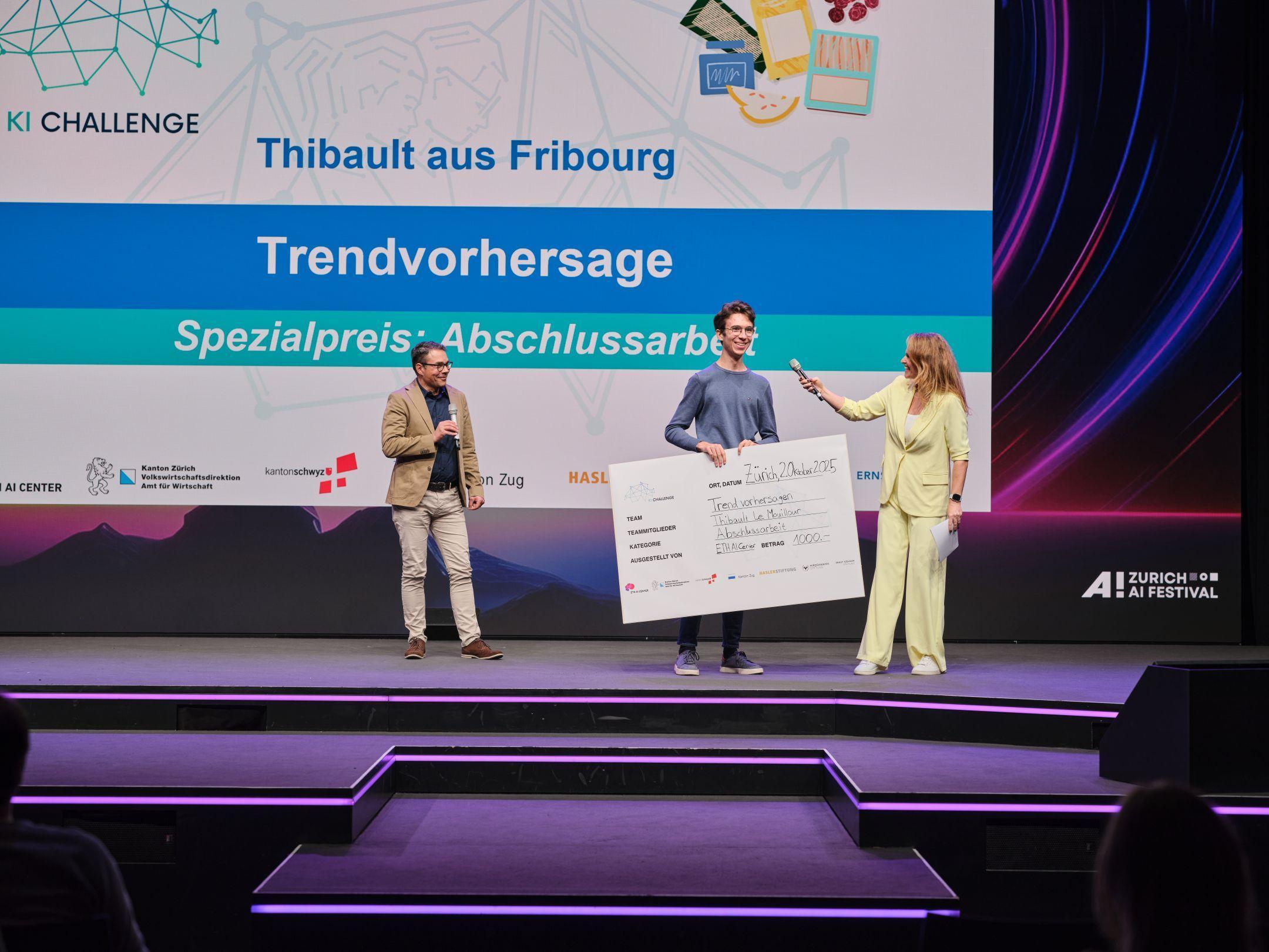 Ein junger Mann nimmt auf der Bühne einen grossen Scheck entgegen, während Markus Müller und Moderatorin Kathrin Hönegger neben ihm stehen.