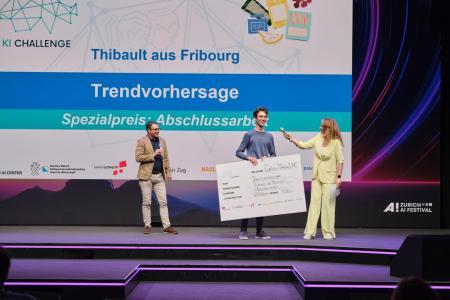 Ein junger Mann nimmt auf der Bühne einen grossen Scheck entgegen, während Markus Müller und Moderatorin Kathrin Hönegger neben ihm stehen.