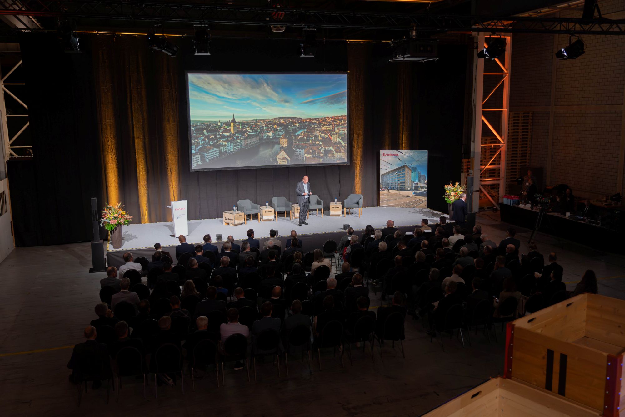 Michael Baumer steht auf der Bühne vor grossem Bildschirm mit Zürich-Panorama und spricht beim Event «Industrie im Dialog» in den Werkhallen von Everllence.