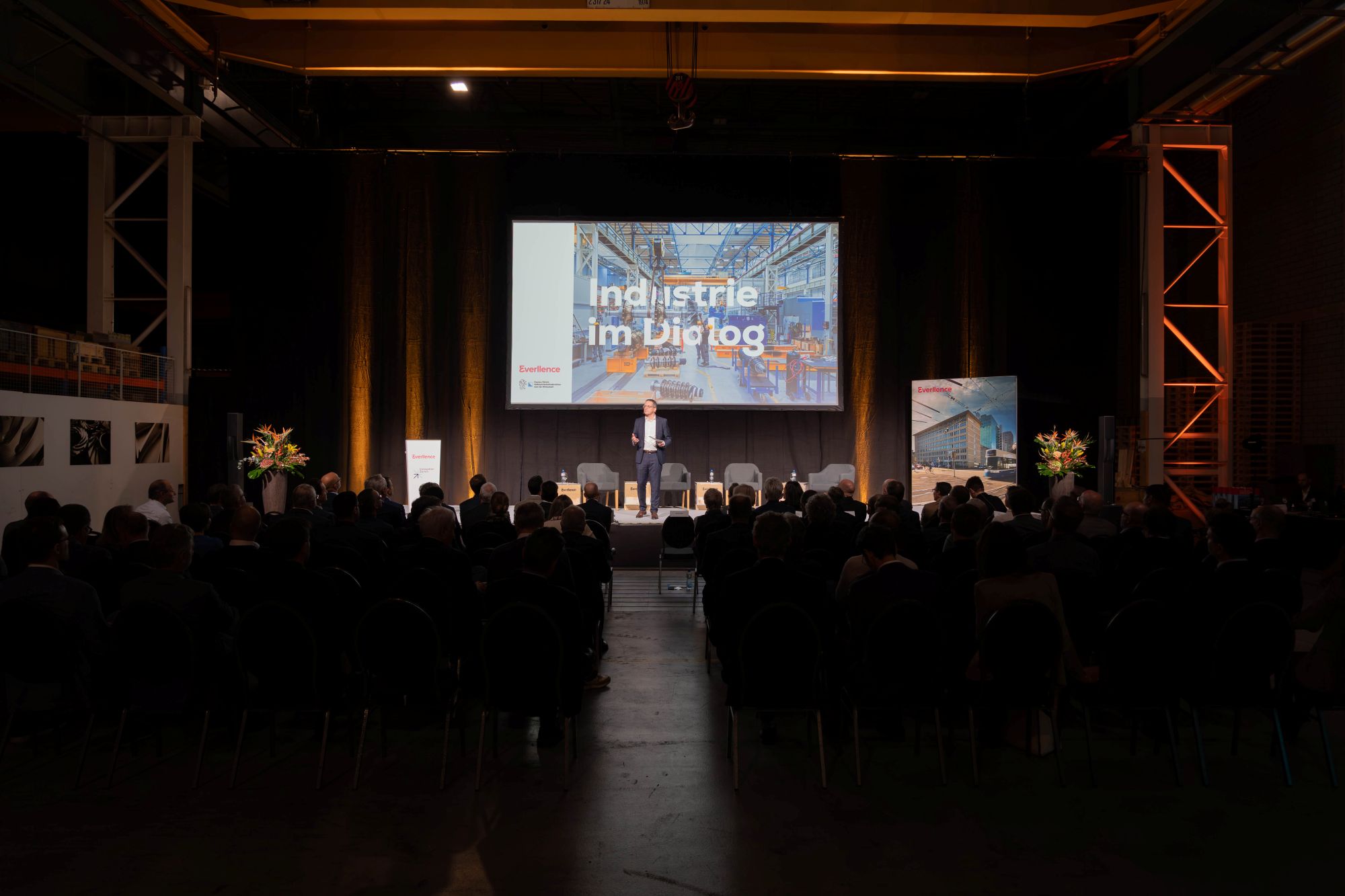 Patrik Meli präsentiert auf der Bühne vor grossem Bildschirm mit dem Titel «Industrie im Dialog» in einer Industriehalle.