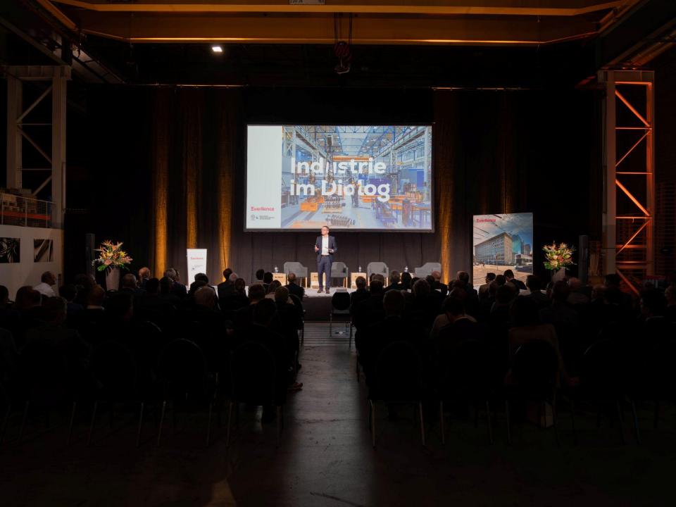 Patrik Meli präsentiert auf der Bühne vor grossem Bildschirm mit dem Titel «Industrie im Dialog» in einer Industriehalle.