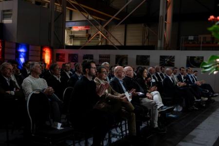 Reihe von Personen sitzt während einer Veranstaltung in einer Industriehalle.