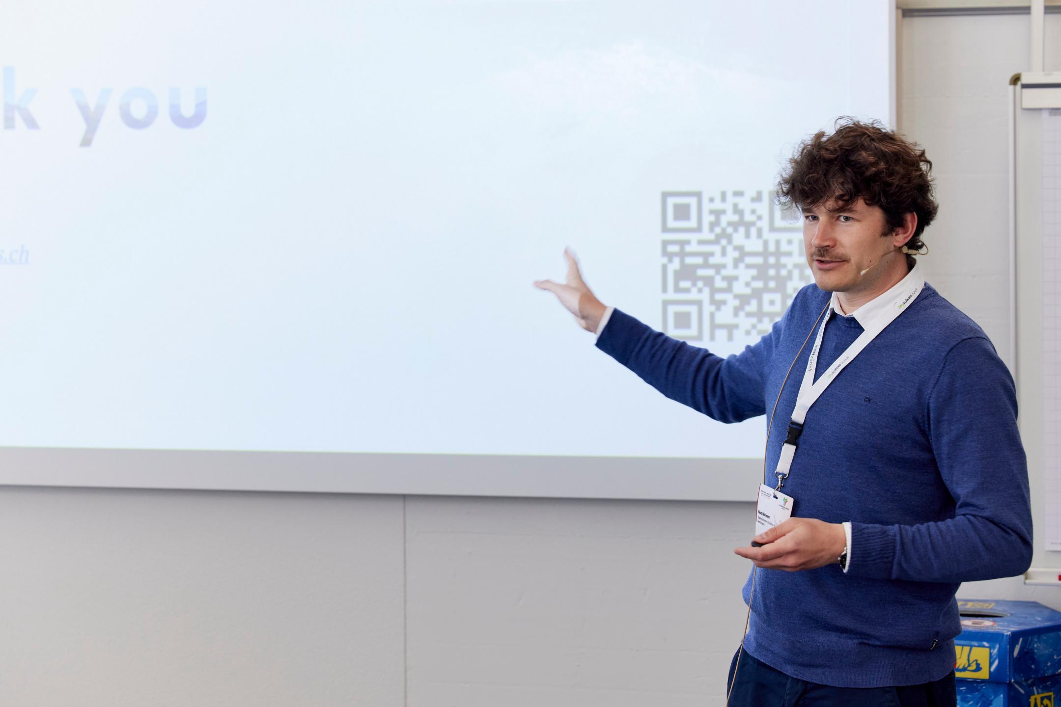 Session Chair Beat Hörmann steht rechts von einer Leinwand und zeigt mit dem rechten Arm auf die Präsentation.