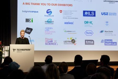 Moderatorin Dr. Anna Erat stellt am Rednerpult auf der Bühne die Ausstellenden vor. Im Hintergrund wird auf der Leinwand eine Folie mit den Logos der Ausstellern gezeigt.