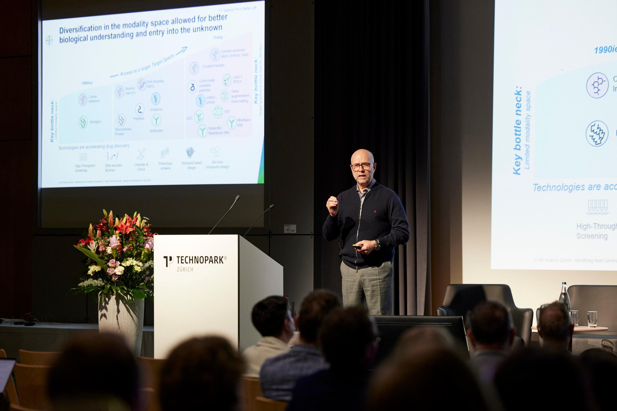 Dr. Christian Rommel hält auf der Bühne des Technoparks Zürich seine Keynote. Im Hintergrund ist eine Folie unscharf zu erkennen.