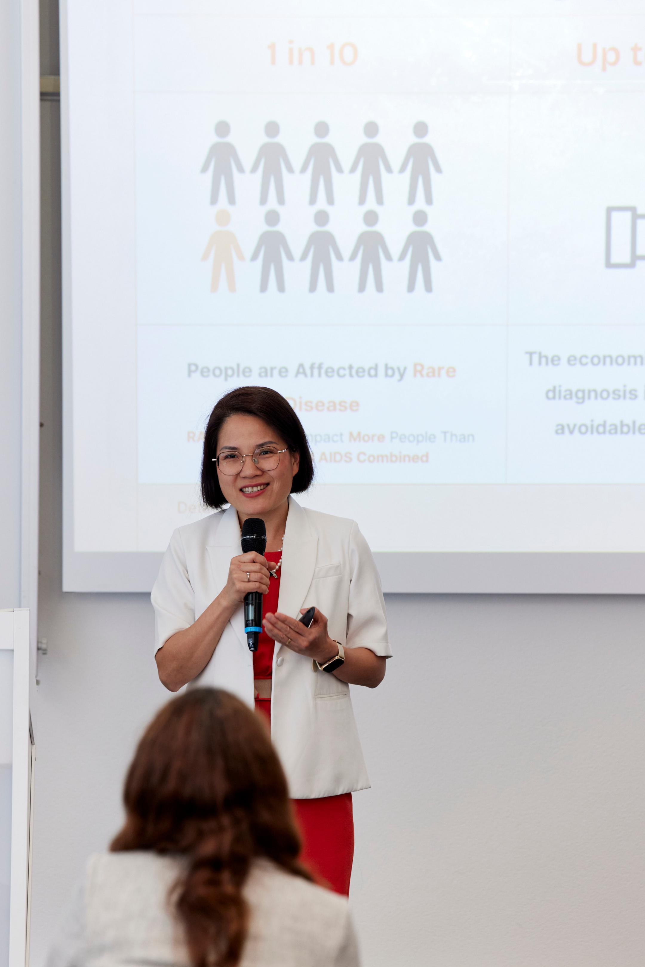 Dr. Quy Vo-Reinhard steht vor einer Leinwand und spricht lächelnd ins Mikrofon. Im Hintergrund sieht man unscharf einen Ausschnitt ihrer Präsentationsfolie.