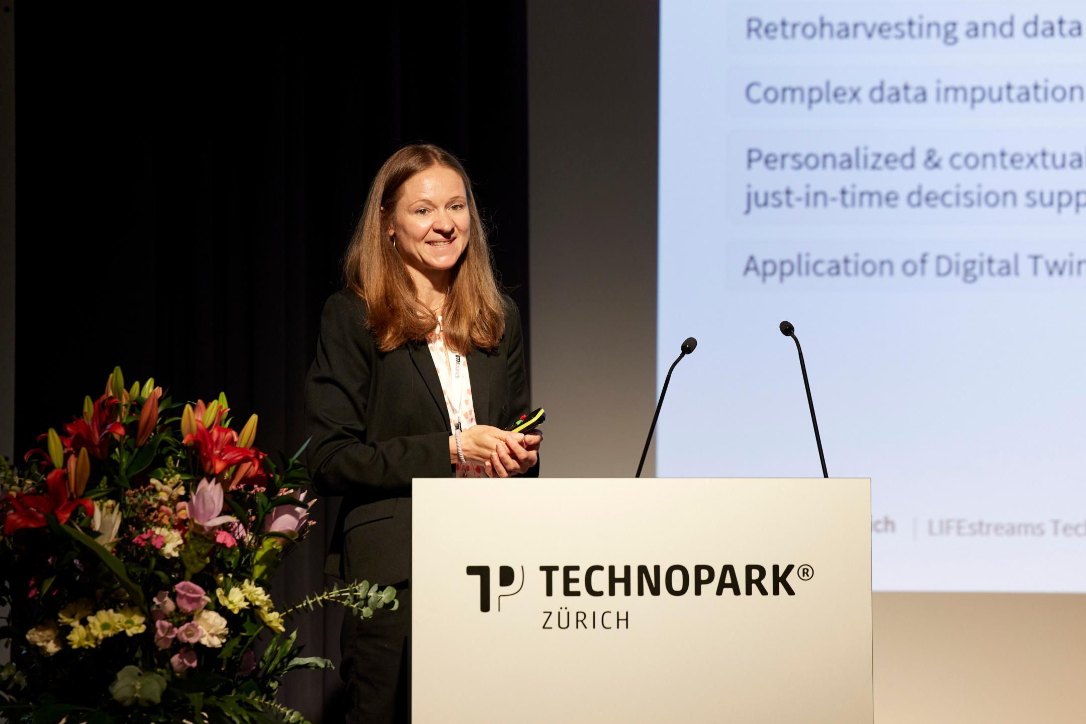 Dr. Susan Mérillat steht hinter dem weissen Rednerpult von Technopark Zürich auf der Bühne und spricht zum Publikum. Im Hintergrund ist ein kleiner Ausschnitt ihrer Präsentation zu sehen.