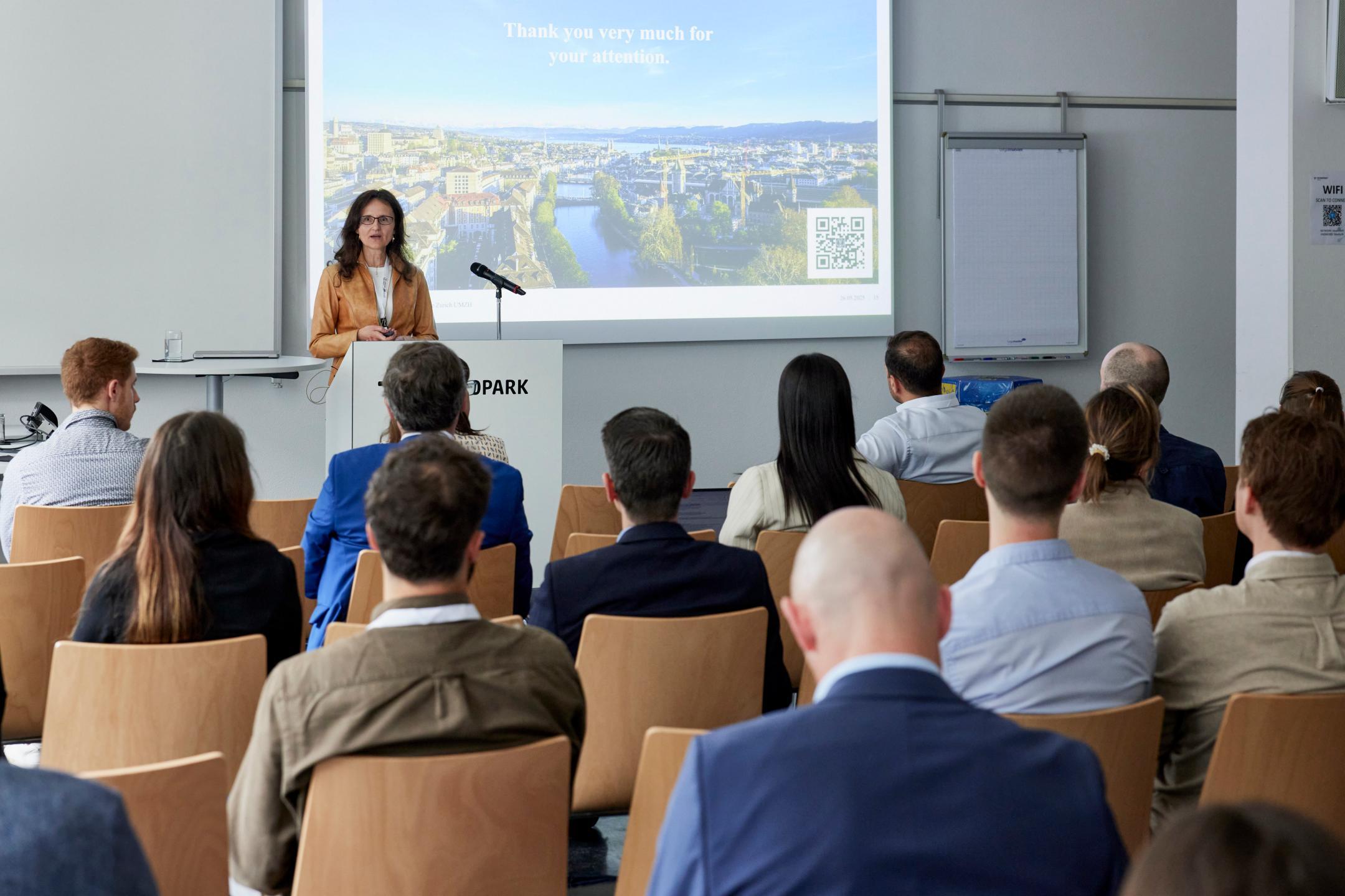 Dr. Suzana Atanasoski steht vor einer Leinwand in einem Meetingraum und spricht zu ihrem Publikum. Auf der Leinwand sieht man unscharf ein Bild einer Stadt mit Bäumen und einem Fluss und See.
