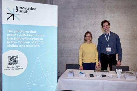 Ein Rollup mit dem Logo von Innovation Zurich und dem Text: The platform that makes collaborations in the field of innovation in the Canton Zurich visible and possible steht links vom Ausstellertisch von Innovation Zurich. Hinter dem Tisch stehen lächelnd eine Frau und ein Mann.