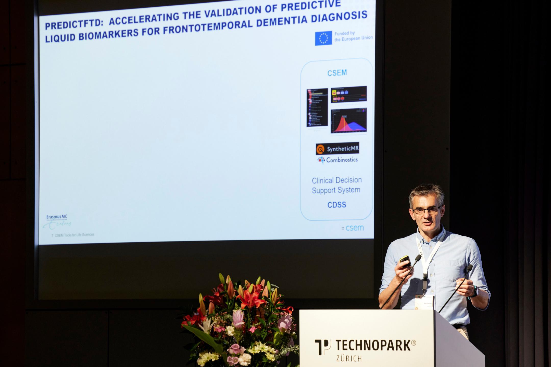 Session Speaker Marc Lamarin steht rechts auf einer Bühne hinter einem weissen Rednerpult und spricht. Hinter ihm ist eine Präsentationsfolie zu sehen.