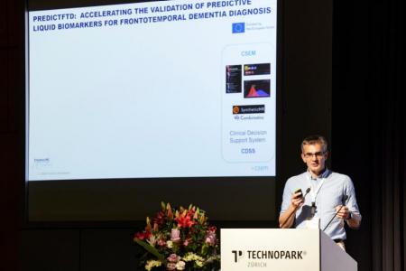 Session Speaker Marc Lamarin steht rechts auf einer Bühne hinter einem weissen Rednerpult und spricht. Hinter ihm ist eine Präsentationsfolie zu sehen.