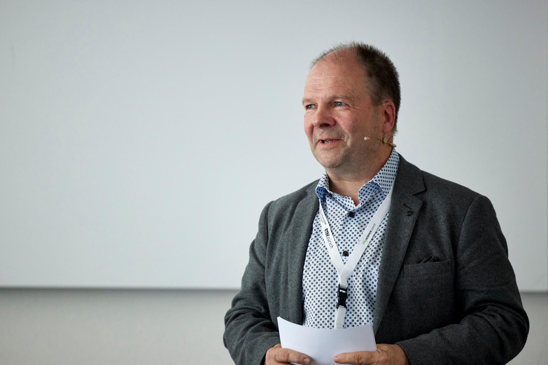 Session Chair Michael J. Flynn ist stehend in Grossaufnahme zu sehen. Er hält ein Blatt Papier und spricht zu seinem Publikum.