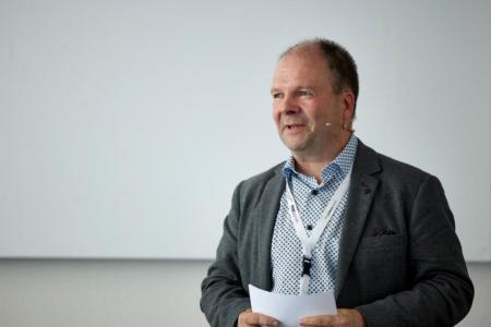 Session Chair Michael J. Flynn ist stehend in Grossaufnahme zu sehen. Er hält ein Blatt Papier und spricht zu seinem Publikum.