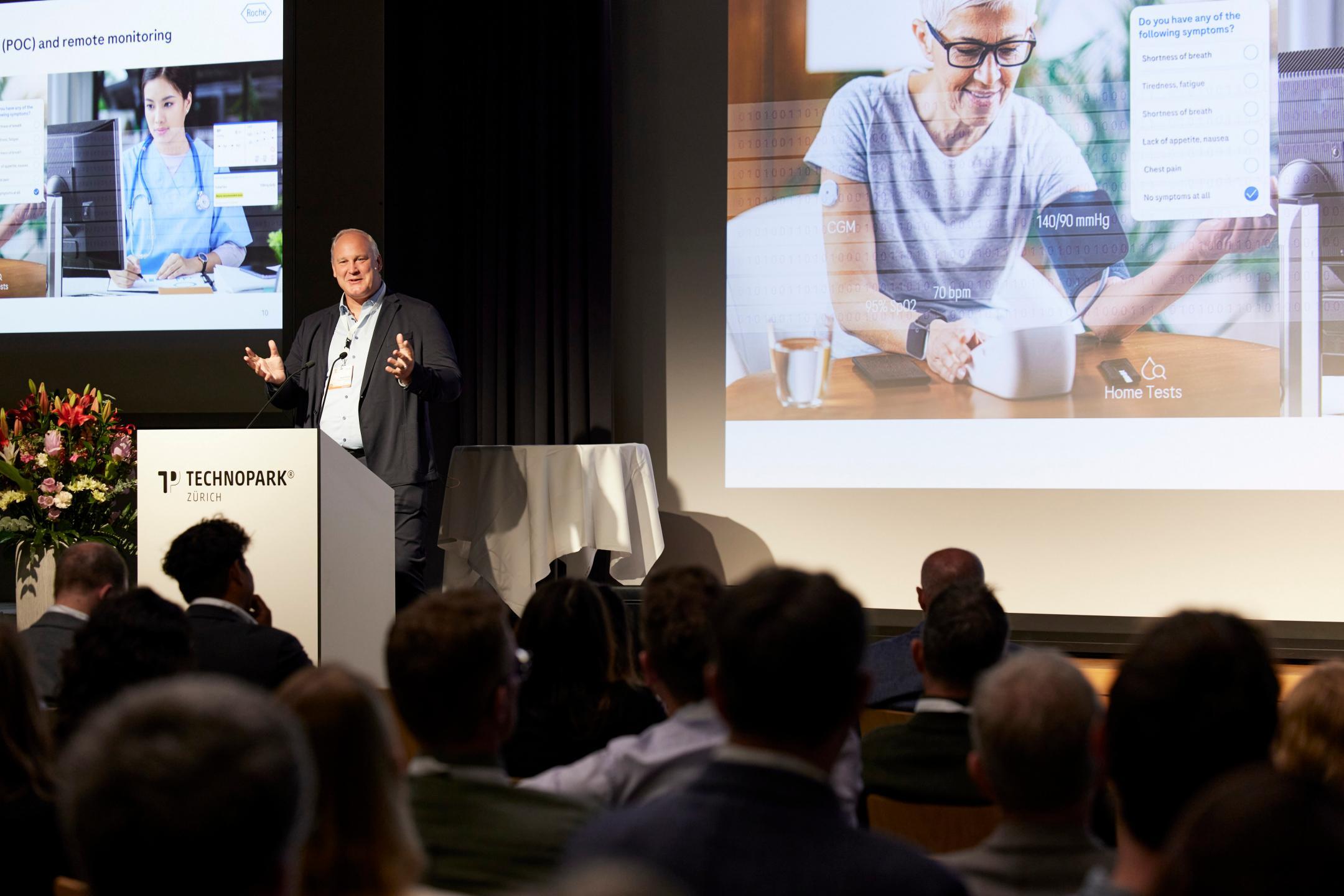 Moritz Hartmann steht als Keynote-Speaker auf der grossen Bühne und spricht zu den Teilnehmenden. Links und rechts von ihm sind zwei Präsentationsfolien zu sehen. Links mit einer jungen Ärztin vor einem Computer und links eine ältere Frau vor einem Pulsmessgerät auf einem Tisch.