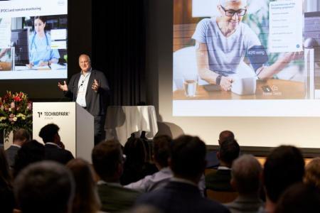 Moritz Hartmann steht als Keynote-Speaker auf der grossen Bühne und spricht zu den Teilnehmenden. Links und rechts von ihm sind zwei Präsentationsfolien zu sehen. Links mit einer jungen Ärztin vor einem Computer und links eine ältere Frau vor einem Pulsmessgerät auf einem Tisch.