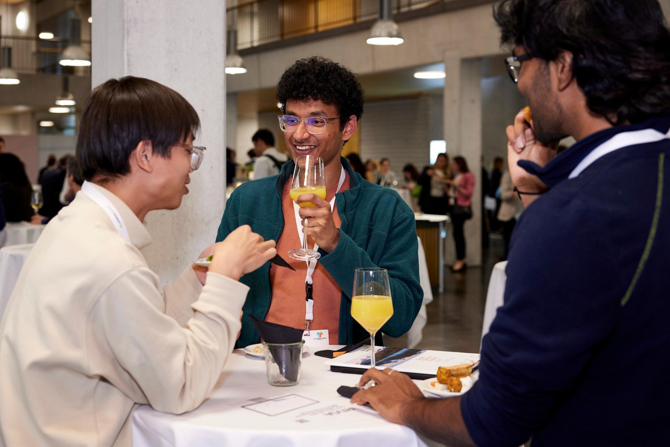 Drei Männer stehen um einen Stehtisch und sprechen miteinander. Zwischen Ihnen stehen Gebäck und Gläser mit Orangensaft.