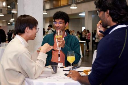 Drei Männer stehen um einen Stehtisch und sprechen miteinander. Zwischen Ihnen stehen Gebäck und Gläser mit Orangensaft.