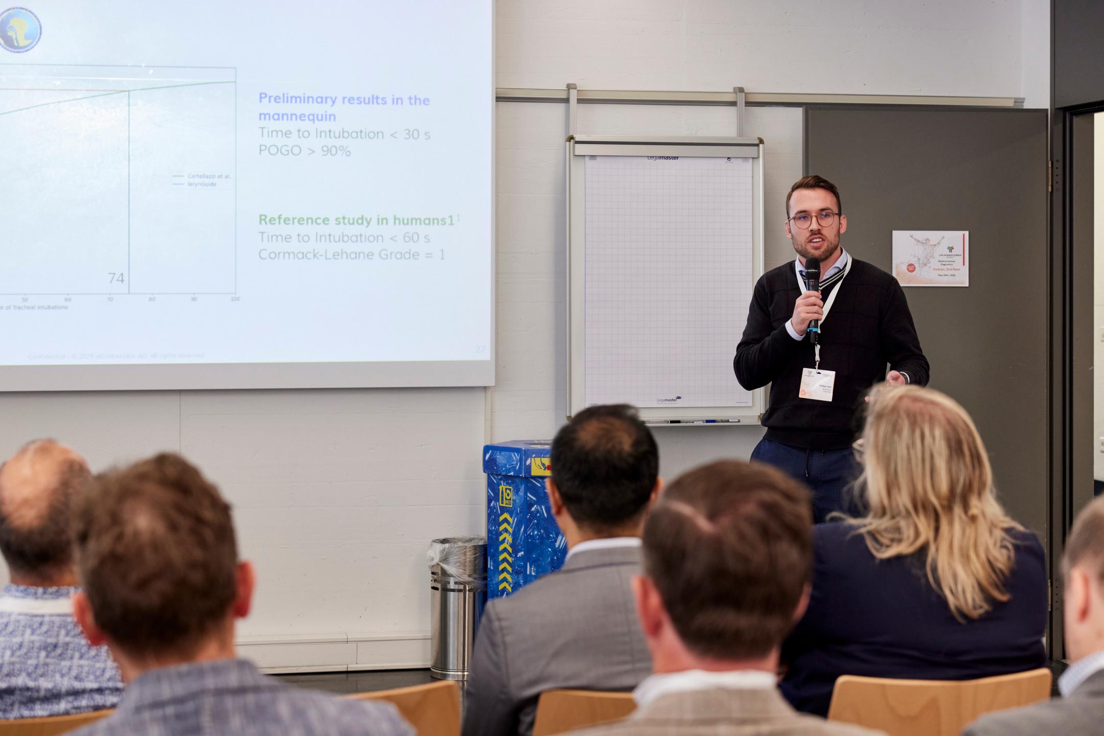 Philipp Ganz spricht zum Publikum in einem Meetingraum. Hinter ihm ist unscharf eine Präsentationsfolie zu sehen und ein leerer Flipchart.