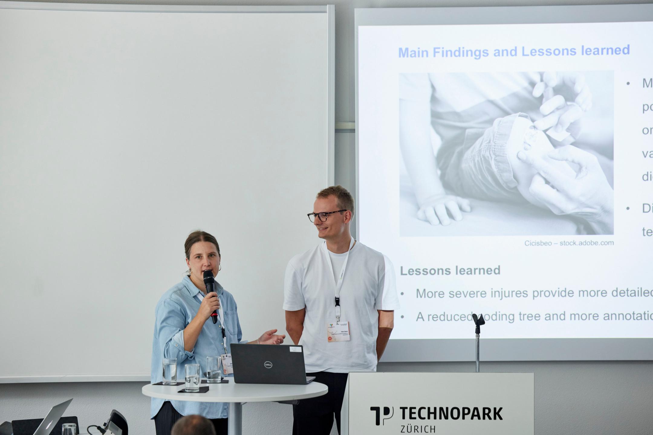 Sonja Feer steht linkd von Felix Saaro in einem Sitzungszimmer. Sie hält das Mikrofon und spricht zum Publikum. Felix Saaro lächelt sie an.