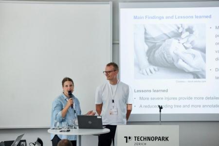 Sonja Feer steht linkd von Felix Saaro in einem Sitzungszimmer. Sie hält das Mikrofon und spricht zum Publikum. Felix Saaro lächelt sie an.