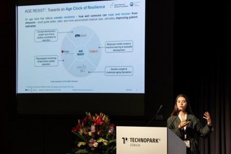Vanessa Matos steht links auf der grossen Bühne und erklärt mit dem Einsatz ihrer Hände ihre Folie die links oben hinter hier zu sehen ist.