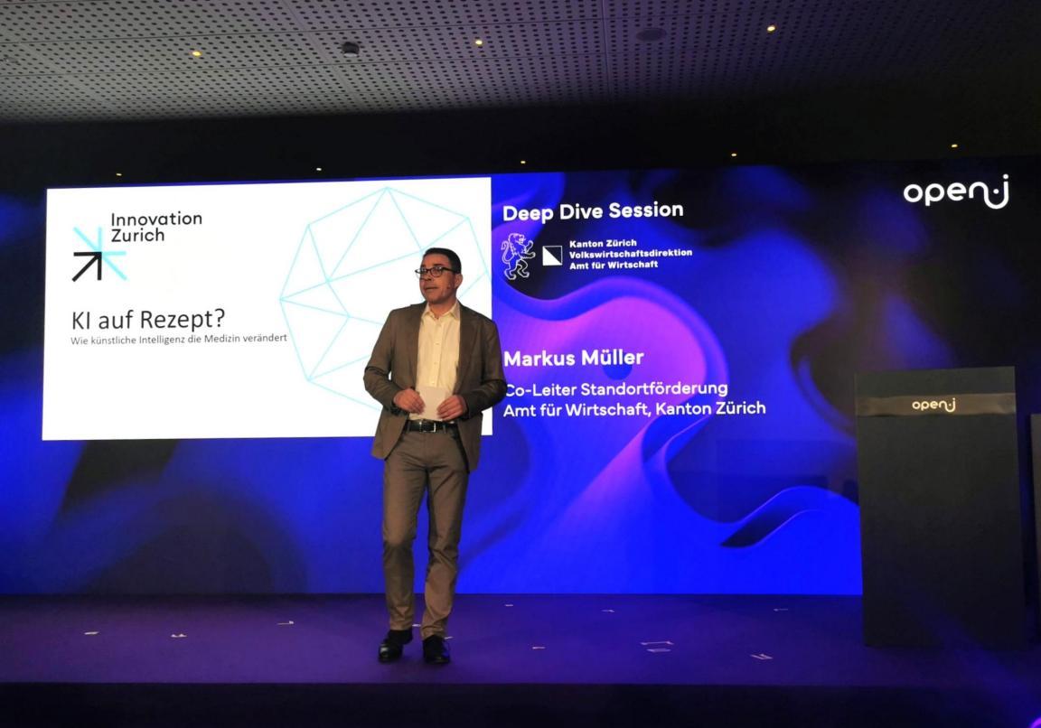 Markus Müller steht auf der Bühne der Deep Dive Session «KI auf Rezept?» vor einer Präsentationswand mit dem Logo des Kantons Zürich und open-i.