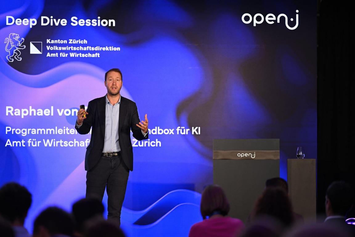 Raphael von Thiessen spricht auf der Bühne der Deep Dive Session «KI auf Rezept?» bei der open-i Innovationskonferenz in Zürich vor einer blauen Präsentationswand mit dem Logo des Kantons Zürich und open-i.