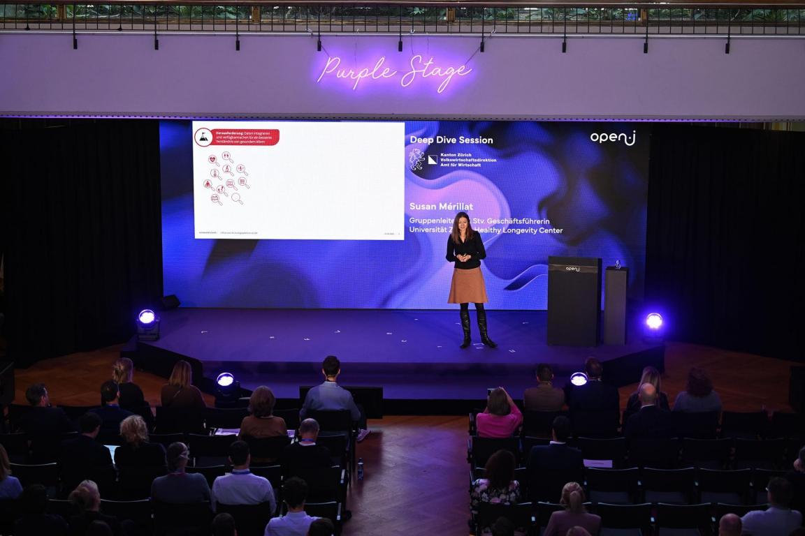 Susan Merillat spricht auf der Purple Stage der open-i Konferenz vor einer grossen Leinwand mit Präsentationsinhalten und dem Titel «Deep Dive Session».