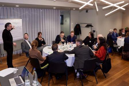 Mehrere Teilnehmende sitzen an einem runden Tisch und diskutieren, während ein Moderator am Flipchart steht.