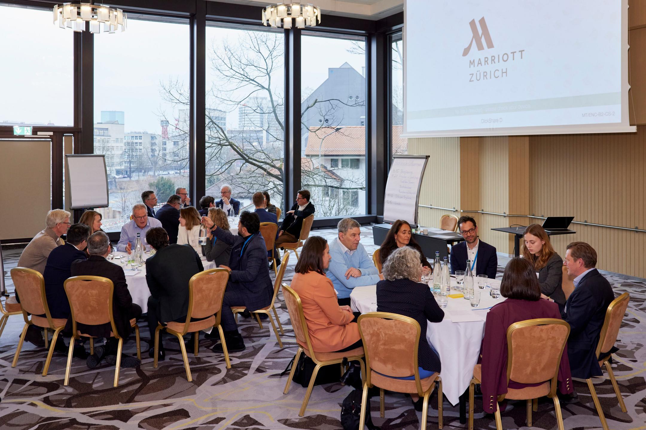 Mehrere Workshopgruppen arbeiten parallel an runden Tischen in einem grossen Veranstaltungsraum.