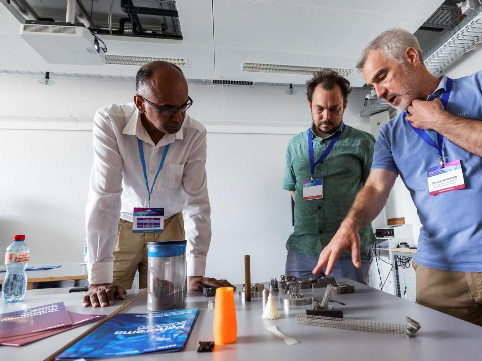 Drei Workshopteilnehmende stehen um einen Tisch mit 3D-Drucker-Produkten und diskutieren über diese