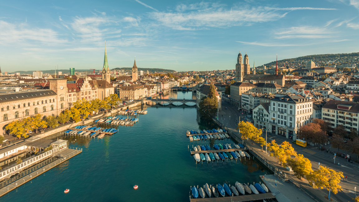 Vogelperspektive auf die Limmat in Zürich bei Sonnenschein, flankiert von den markanten Kirchtürmen des Grossmünsters und der Fraumünster-Kirche sowie zahlreichen Booten an den Stegen.