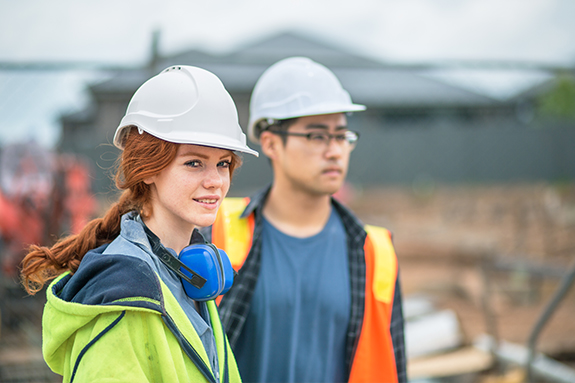 Eine Frau und ein Mann, mit Helm auf dem Kopf, stehen auf einer Baustelle.
