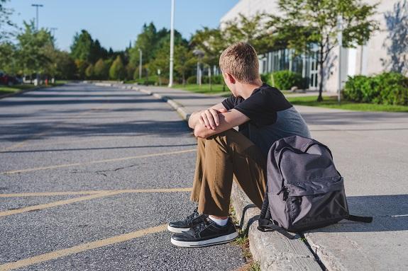 Teenagerjunge sitzt verlassen am Strassenrand, daneben steht sein Schul-Rucksack.