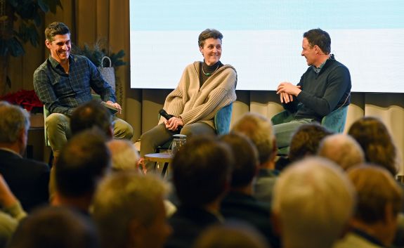 Drei Personen &ndash; zwei M&auml;nner und eine Frau &ndash; sitzen l&auml;chelnd auf St&uuml;hlen vor einer hellen Leinwand und einem Publikum, das im Vordergrund unscharf zu sehen ist.