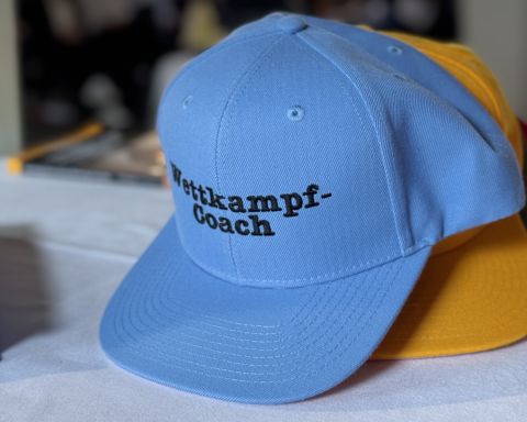 Hellblaue Baseballkappe mit der Bestickung &bdquo;Wettkampf-Coach&ldquo; in Schwarz, dahinter liegt eine gelbe Kappe.