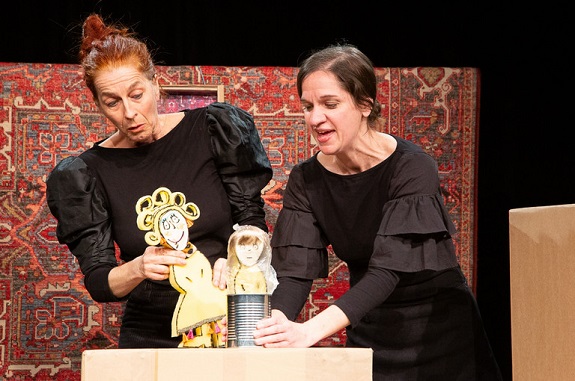 Zwei Frauen in dunklen Kleidern mit zwei Kartonpuppen. Die eine Puppe guckt aus einer Konservenbüchse