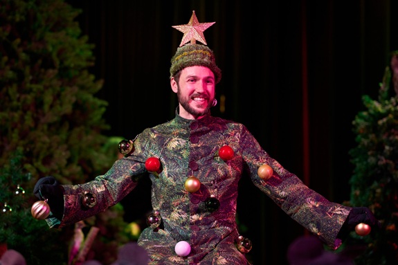 Das Bild zeigt eine Person in einem Weihnachtsbaum-Kost&uuml;m mit bunten Kugeln und einem Stern auf dem Kopf. Im Hintergrund sind B&auml;ume zu sehen.