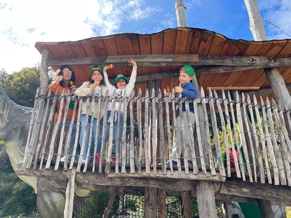 Das Bild zeigt vier Kinder auf einer h&ouml;lzernen Plattform eines Baumhausbaus. Drei tragen gr&uuml;ne Stirnb&auml;nder. Links ist ein Dinosauriermodell zu sehen, im Hintergrund B&auml;ume und ein teils bew&ouml;lkter Himmel.