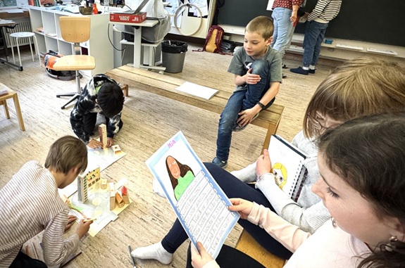 Das Bild zeigt ein Klassenzimmer mit mehreren Kindern, die an verschiedenen Aktivit&auml;ten beteiligt sind &ndash; einige sitzen auf dem Boden und arbeiten, andere stehen oder sitzen nahe der Tafel.
